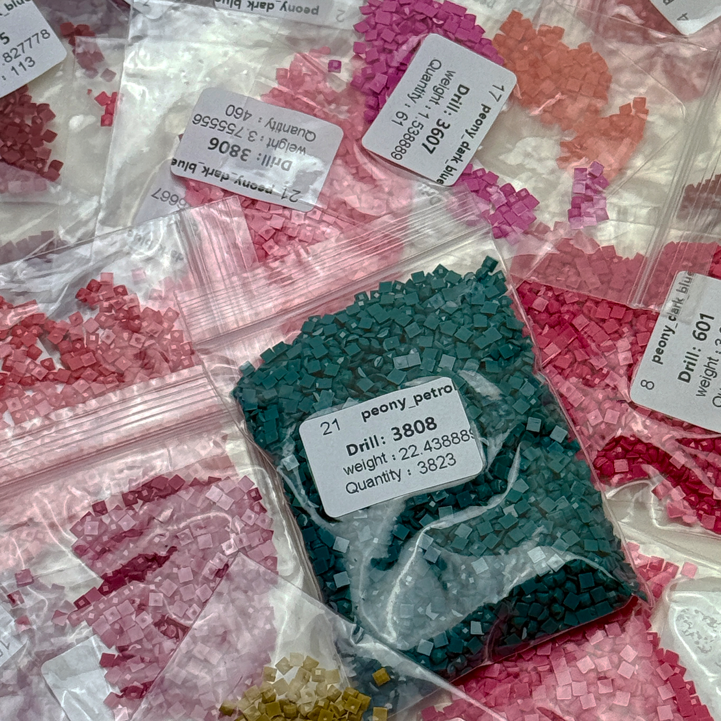 Foto mit Diamond-Painting-Steinchen des Micromosaic Motivs 'peony-petrol', abgepackt in Zipper-Tütchen und mit Etiketten versehen, auf denen die Farbnummern gekennzeichnet sind.