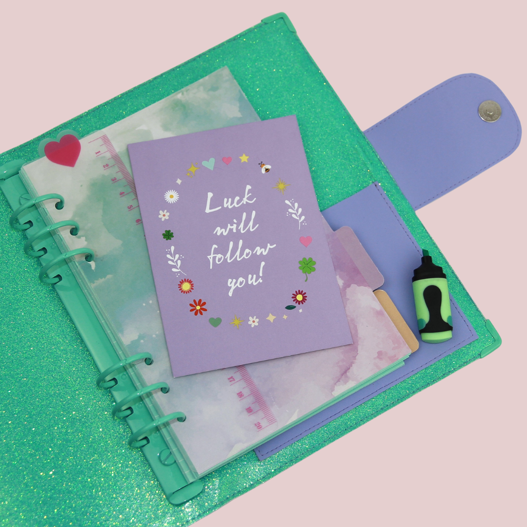 Nahaufnahme des geöffneten A5-Planers mit grün glitzernder Innenseite, Ringmechanik, Steckfächern und Einlagen; in der Mitte liegt eine lilafarbene Karte mit der Aufschrift „Luck will follow you“.